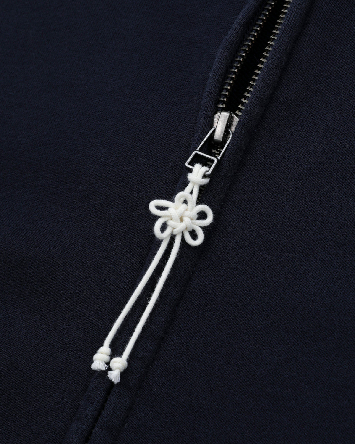 Close-up of the NAVY WASH FLOWER KNOT ZIP UP from Sundae School, made from navy blue cotton and finished with a silver zipper. The zipper pull is adorned with a decorative white Maehwa knot in a floral shape, its two loose ends elegantly dangling below.