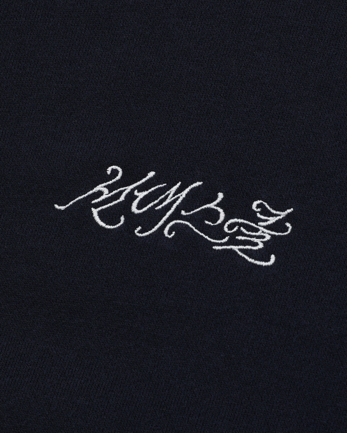 White calligraphic text is embroidered on a dark cotton background, reminiscent of the ornate lettering associated with Sundae School. The script is stylized and intricate, lending an enigmatic appeal to the NAVY WASH FLOWER KNOT ZIP UP hoodie by Sundae School.