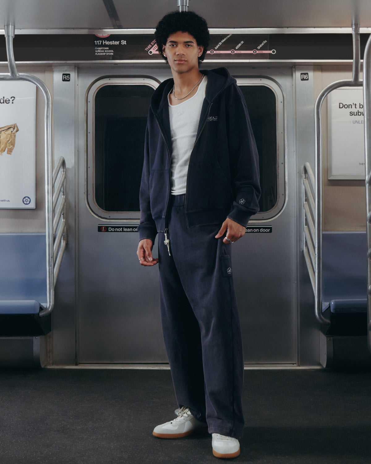 A person wearing the Sundae School NAVY WASH WRAP SWEAT BAJI joggers and white sneakers stands in an empty subway car, facing the camera in front of closed doors with visible signs and handrails.