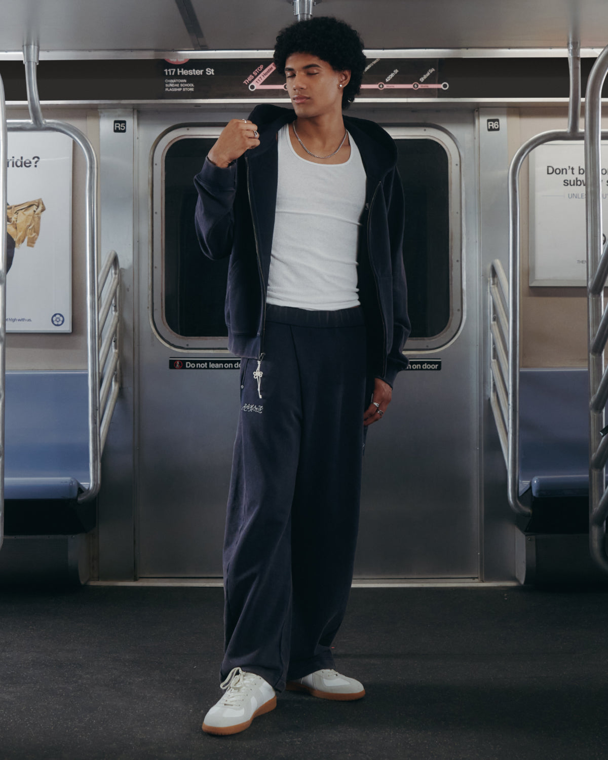Wearing the Sundae School NAVY WASH WRAP SWEAT BAJI and a dark zip-up hoodie over a white tank top, a person stands on a subway train, looking to the side with one hand on their hoodie zipper.