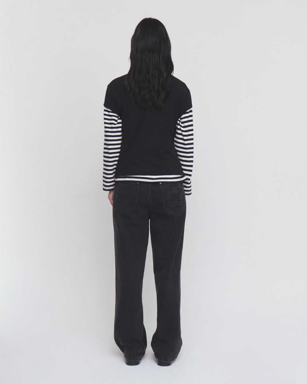 A person stands with their back to the camera against a plain white background, wearing Sundae School’s BLACK WIDOW FRESHMAN FIT DENIM and a black long-sleeve top featuring black and white striped sleeves.