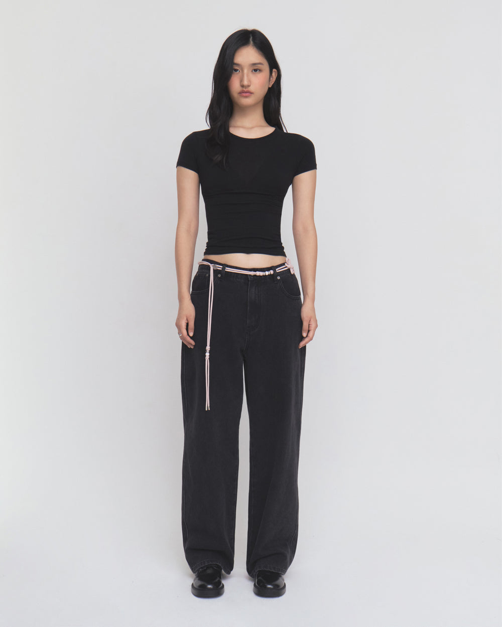 A woman stands against a plain white background, wearing a fitted black short-sleeve top and Sundae School BLACK WIDOW FRESHMAN FIT DENIM jeans styled with a delicate Norigae belt, paired with black shoes. She has long dark hair and a neutral expression.