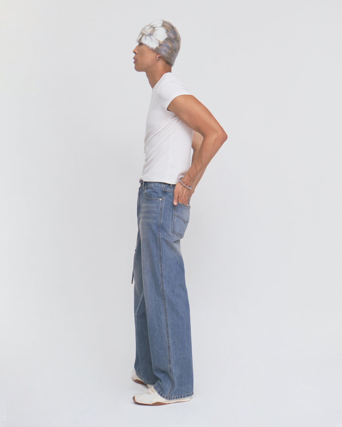 A person stands in profile against a white background, wearing a white t-shirt, Sundae School’s MEDIUM BLUE FRESHMAN FIT DENIM jeans in a wide-leg style, white shoes, and a patterned beanie. Their left hand rests on their hip.