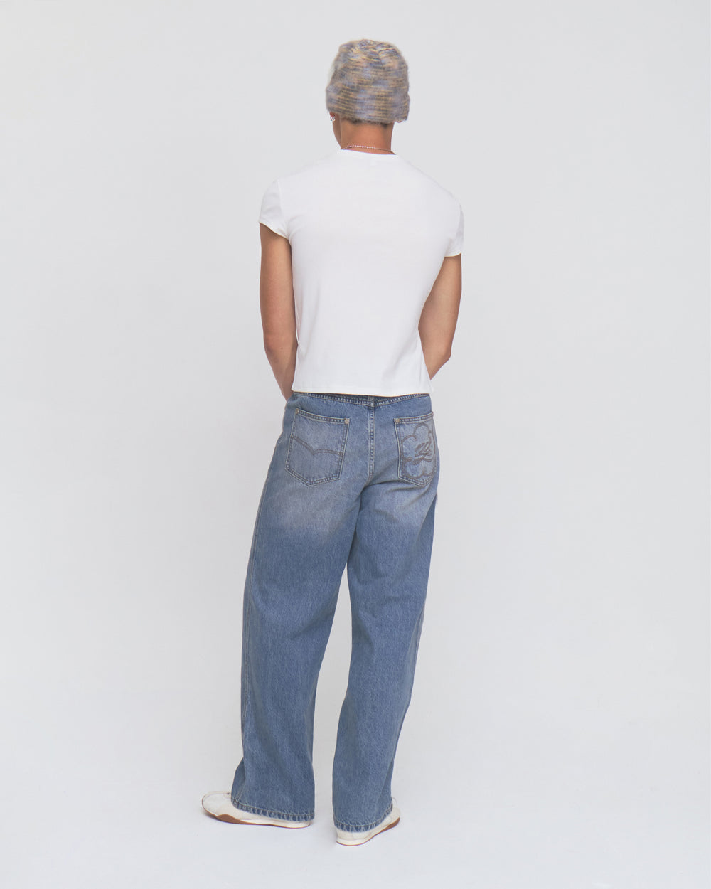 A person stands facing away, wearing a gray beanie, a white short-sleeve shirt, Sundae School’s MEDIUM BLUE FRESHMAN FIT DENIM jeans, and white shoes against a plain light background.