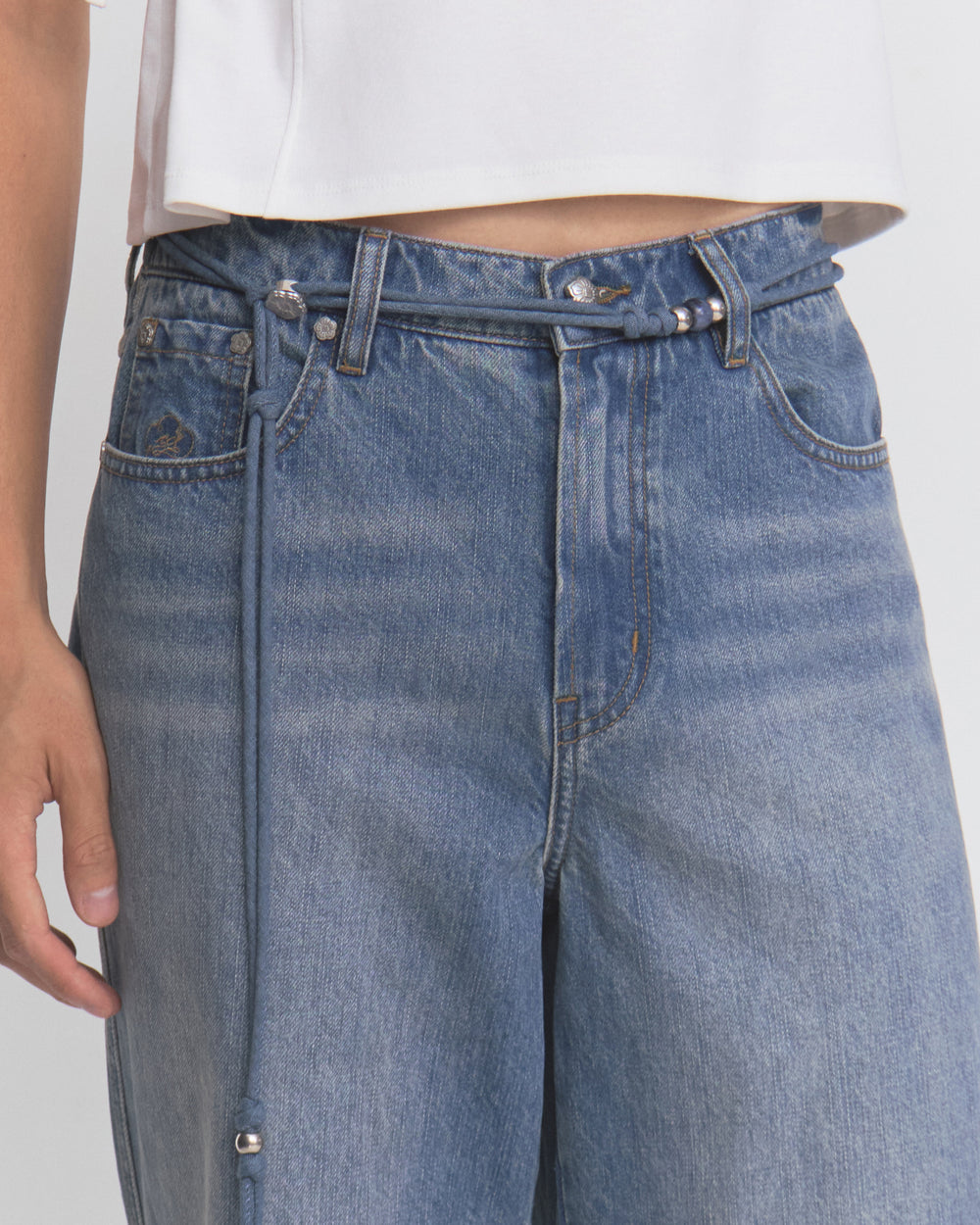 A person wearing Sundae School’s MEDIUM BLUE FRESHMAN FIT DENIM jeans with a thin denim belt and a white cropped top, shown from mid-torso to upper thigh. The person’s left hand is visible by their side.