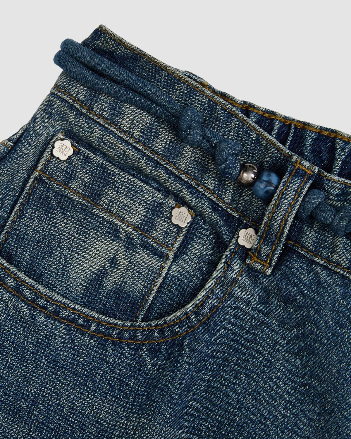 Close-up of Sundae School’s MEDIUM BLUE FRESHMAN FIT DENIM, highlighting relaxed straight-leg jeans with a front pocket, silver rivets, and a braided Norigae belt for unique Korean style against a plain background.