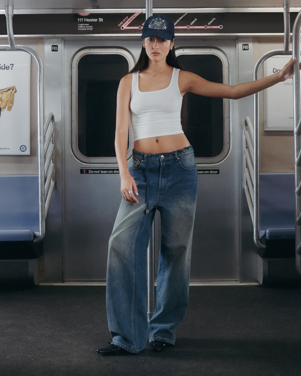 A person wearing a white tank top, Sundae School’s BLUE DREAM FRESHMAN FIT DENIM, and a blue cap stands inside a subway car, holding onto a pole with the subway doors and seats visible in the background.