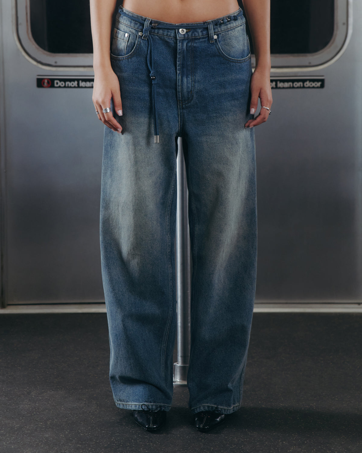 A person wearing Sundae School MEDIUM BLUE FRESHMAN FIT DENIM and black shoes stands in front of a subway door with a 