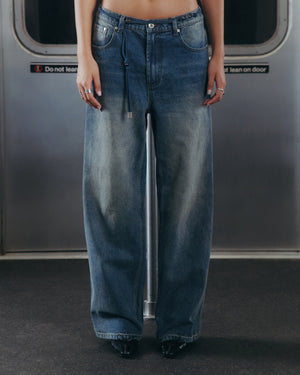 A person wearing Sundae School MEDIUM BLUE FRESHMAN FIT DENIM and black shoes stands in front of a subway door with a "Do not lean on door" sign visible. The waist and ankles are cropped from the image.