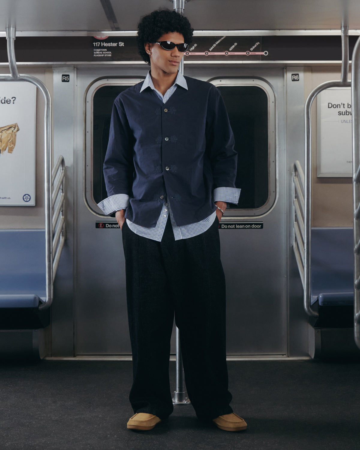Wearing sunglasses and the Sundae School MAEHWA COLLARLESS SHIRT beneath a navy button-up jacket, along with wide-leg pants and tan shoes, a person stands in the middle of an empty subway car.
