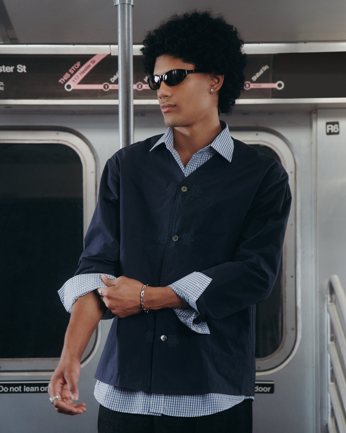 A person with curly hair and sunglasses stands in a subway car, wearing a dark jacket over the MAEHWA COLLARLESS SHIRT by Sundae School. A subway map is visible in the background.