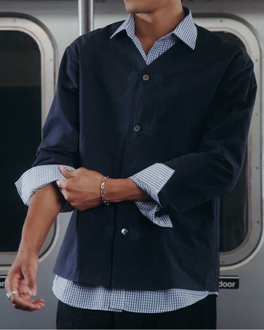 A man wearing a dark navy overshirt over a light blue and white gingham check shirt, standing in front of a train window.