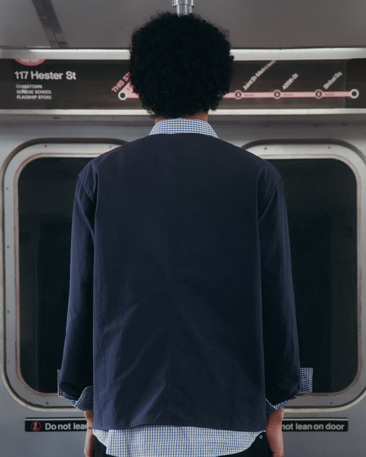 A person with curly hair stands facing a subway door, back to the camera. A subway map and station sign 