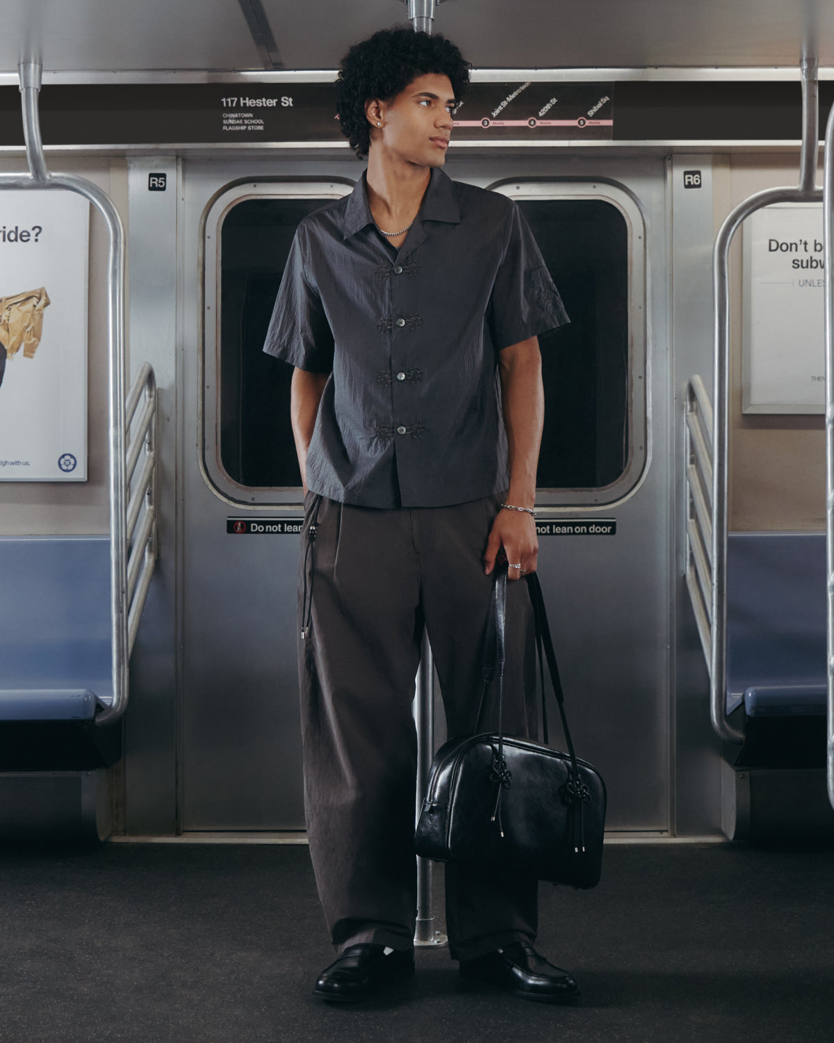 A person with curly hair stands in a well-lit, clean subway car with empty seats, wearing the Sundae School MAEHWA EMBROIDERED SHEER SHIRT and loose pants, while holding a black bag.