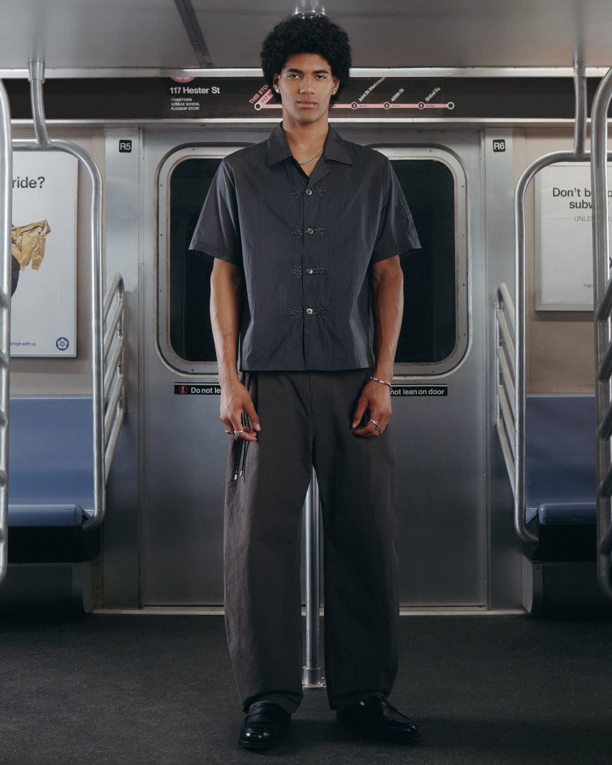 A person with curly hair stands in the middle of a subway car, wearing the Sundae School MAEHWA Embroidered Sheer Shirt paired with loose pants, holding something in their right hand. The subway interior is metallic with empty seats and visible signage.
