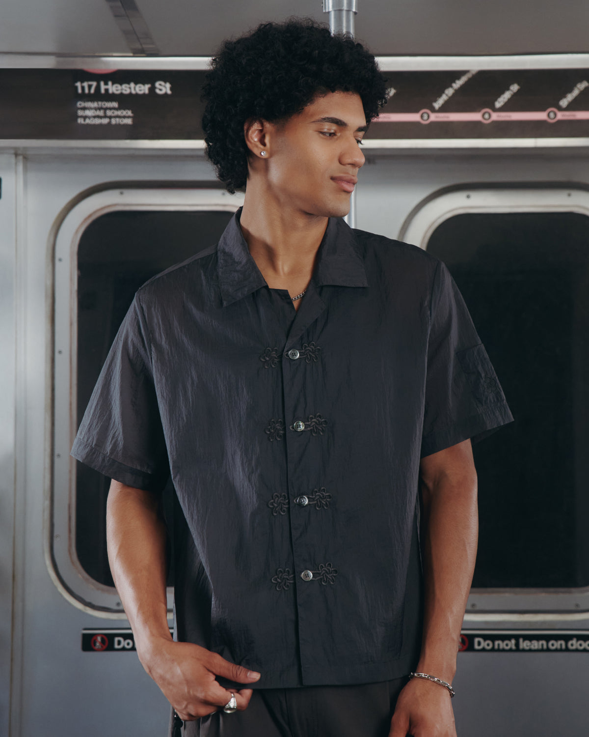 A person with curly hair, dressed in the Sundae School MAEHWA EMBROIDERED SHEER SHIRT, stands inside a subway train near the doors. A sign above reads 117 Hester St. The person looks to the side with one hand in their pocket.