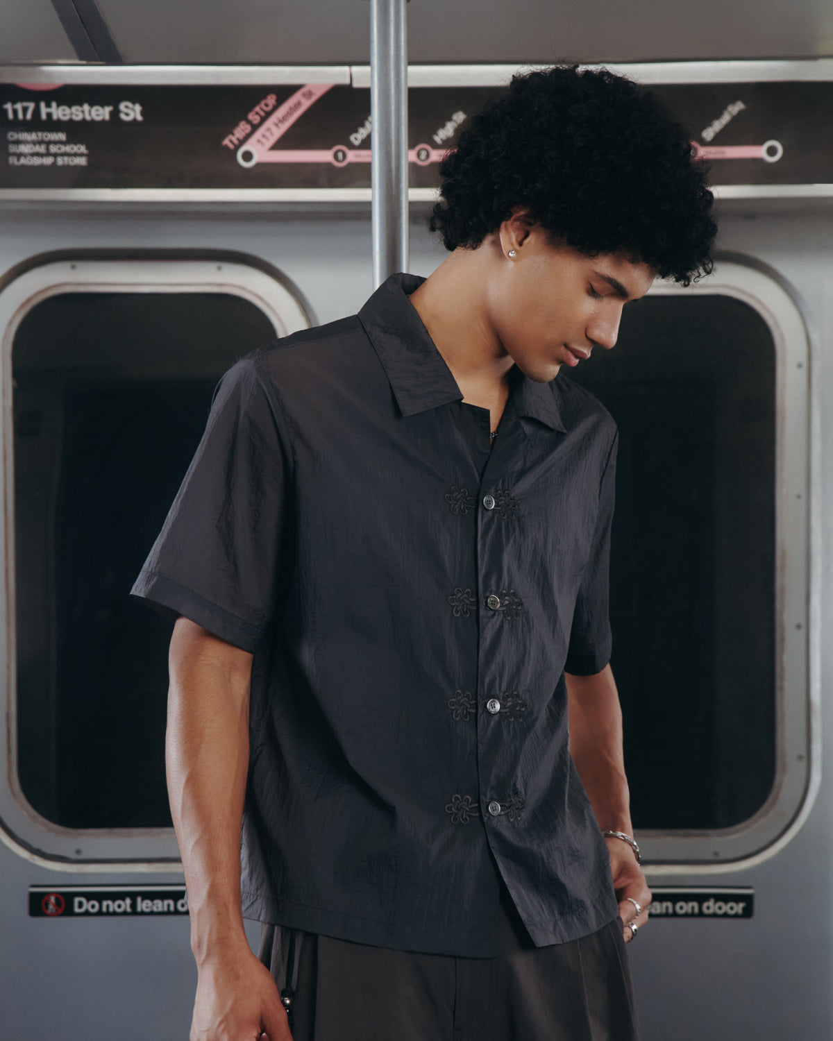 Wearing the Sundae School MAEHWA EMBROIDERED SHEER SHIRT, a person with curly hair stands on a subway train in front of windows and a sign that reads Hester St., looking down with a calm expression.