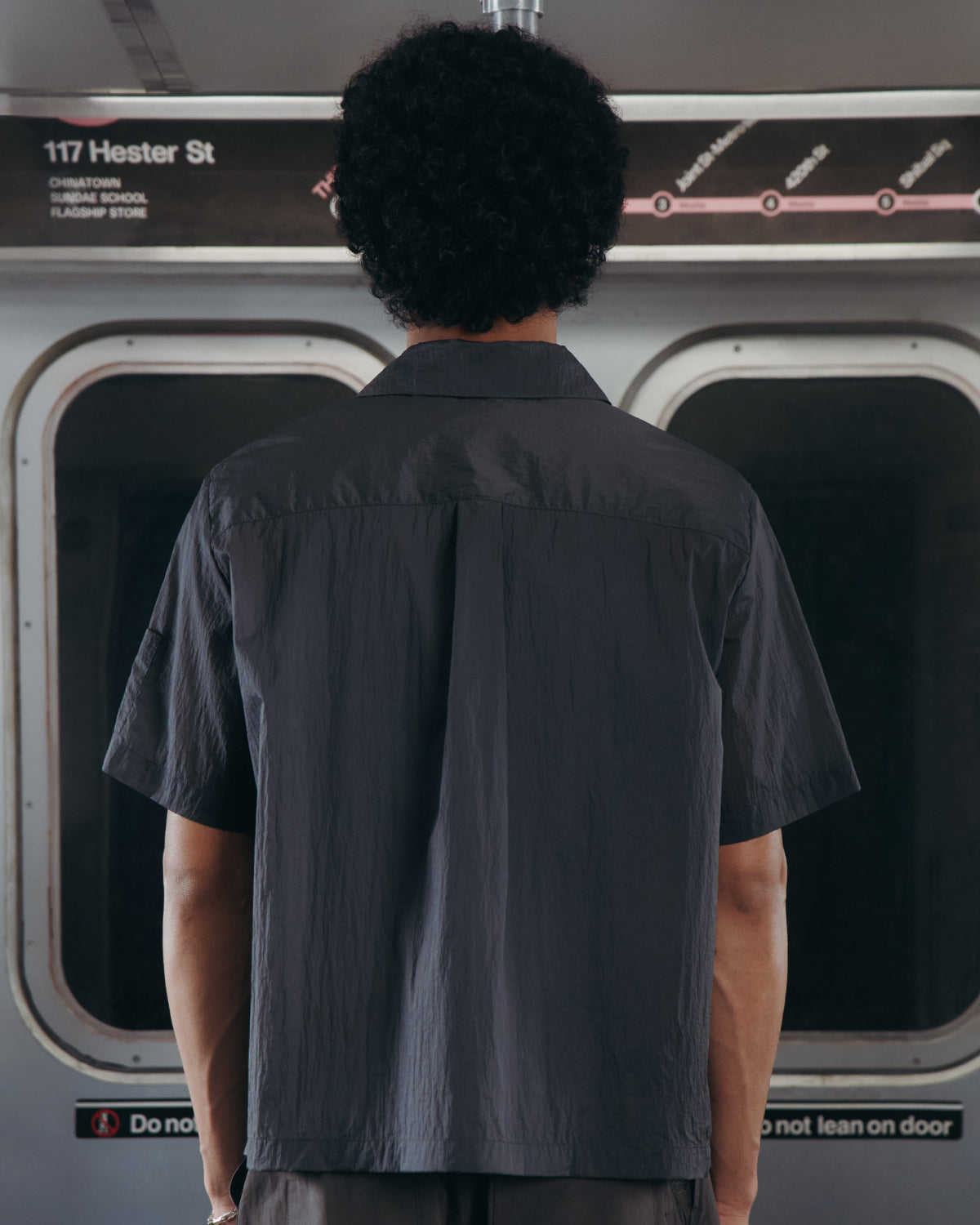 Wearing the Sundae School MAEHWA EMBROIDERED SHEER SHIRT, a person with curly hair stands with their back to the camera inside a subway train near a sign reading 117 Hester St.