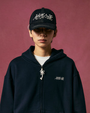 A person wearing a black hoodie and a Sundae School INDIGO FLOWER KNOT CAP stands against a plain maroon background, looking directly at the camera.