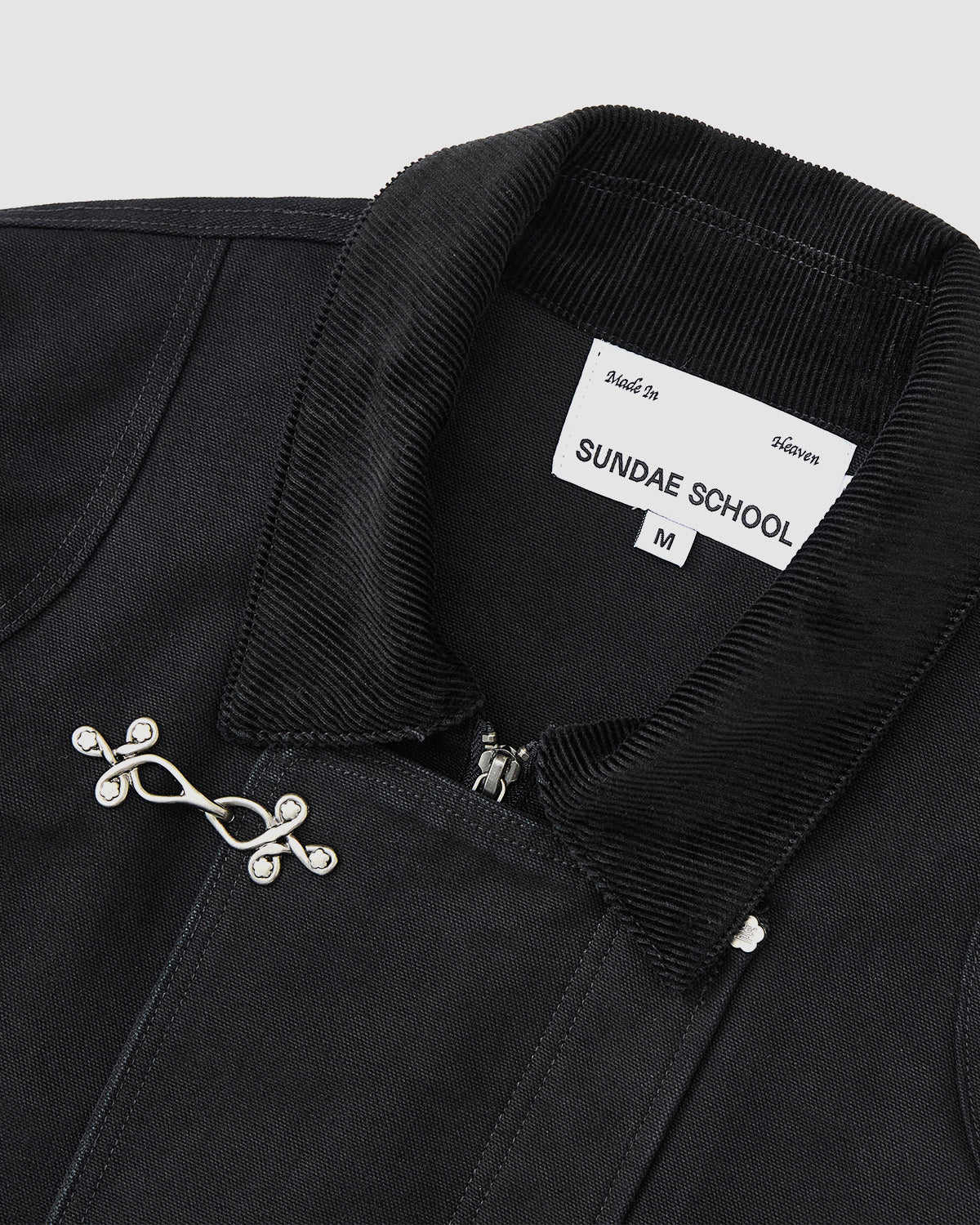 Close-up of the [PRE-ORDER] BLACK FIREMAN CLASP JACKET by Sundae School, featuring a black cotton body, corduroy collar, Mochi Floral clasp, and a label displaying Sundae School, size M, and 