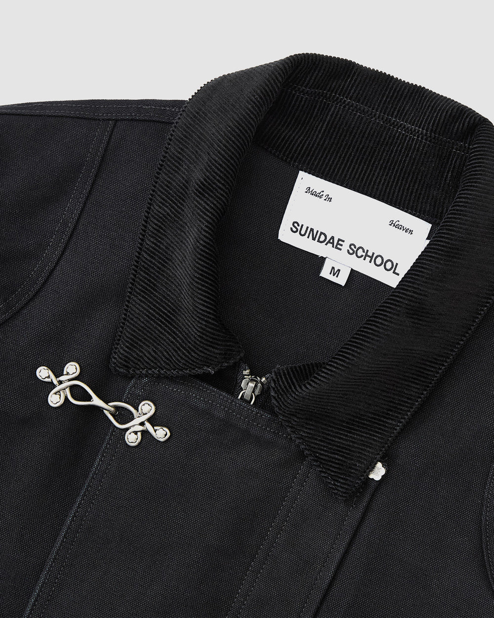 Close-up of the [PRE-ORDER] BLACK FIREMAN CLASP JACKET by Sundae School, featuring a black cotton body, corduroy collar, Mochi Floral clasp, and a label displaying Sundae School, size M, and "Made in Heaven.