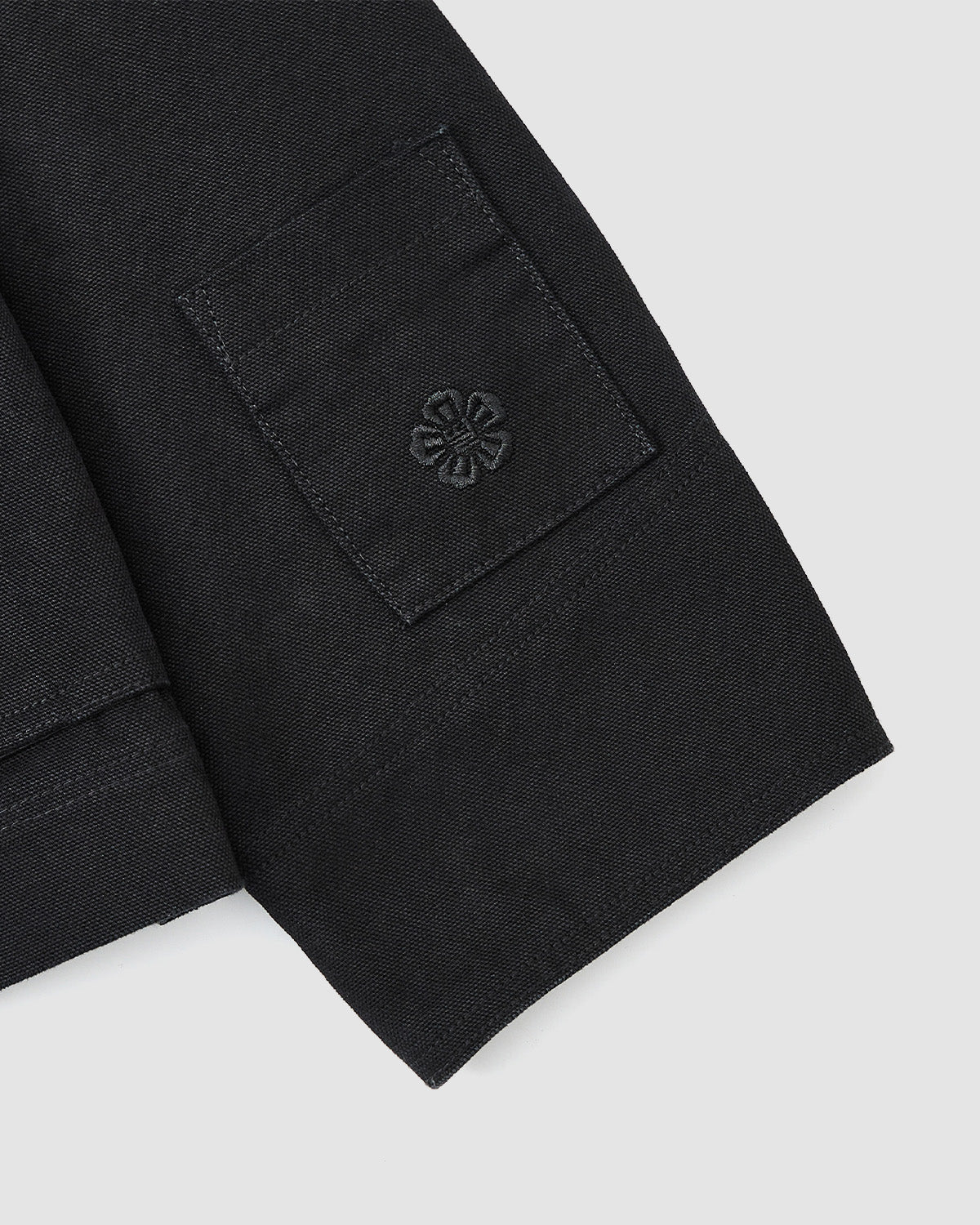 A close-up of the Sundae School [PRE-ORDER] BLACK FIREMAN CLASP JACKET sleeve shows a rectangular patch with a black embroidered flower emblem, set against a light gray background.