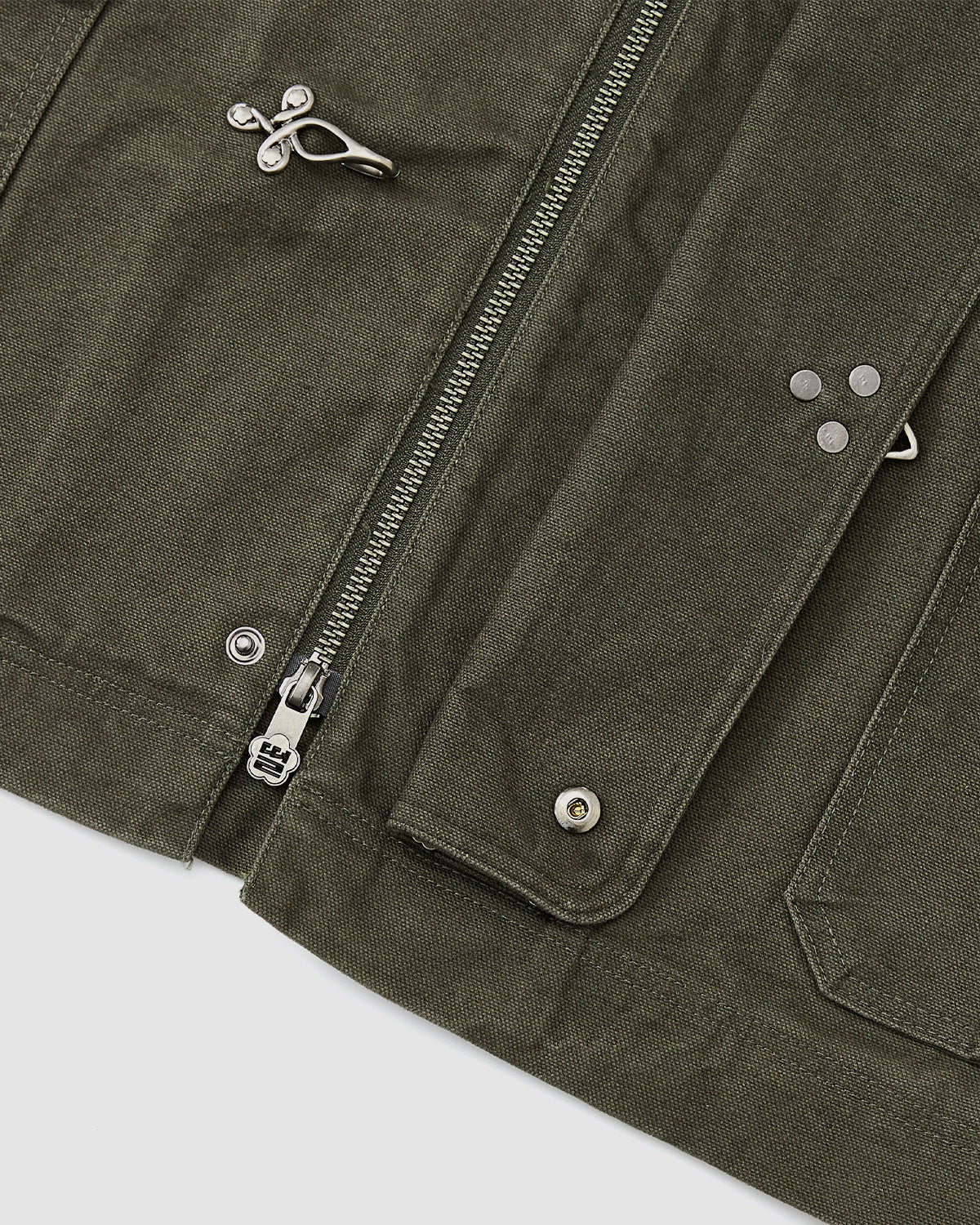 Close-up of the [PRE-ORDER] OLIVE FIREMAN CLASP JACKET by Sundae School, a vintage-inspired cotton workwear piece in olive green with a sturdy textured fabric, silver zipper, metal snap buttons, and decorative silver pins on the front pocket and flap.