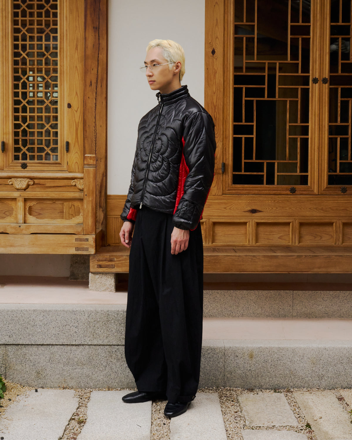 A person with blond hair and glasses wears the Sundae School BLACK RED MOCHI SPIRAL PUFFER, loose black pants, and black shoes as they stand on stone steps before a wooden building with intricate window designs.