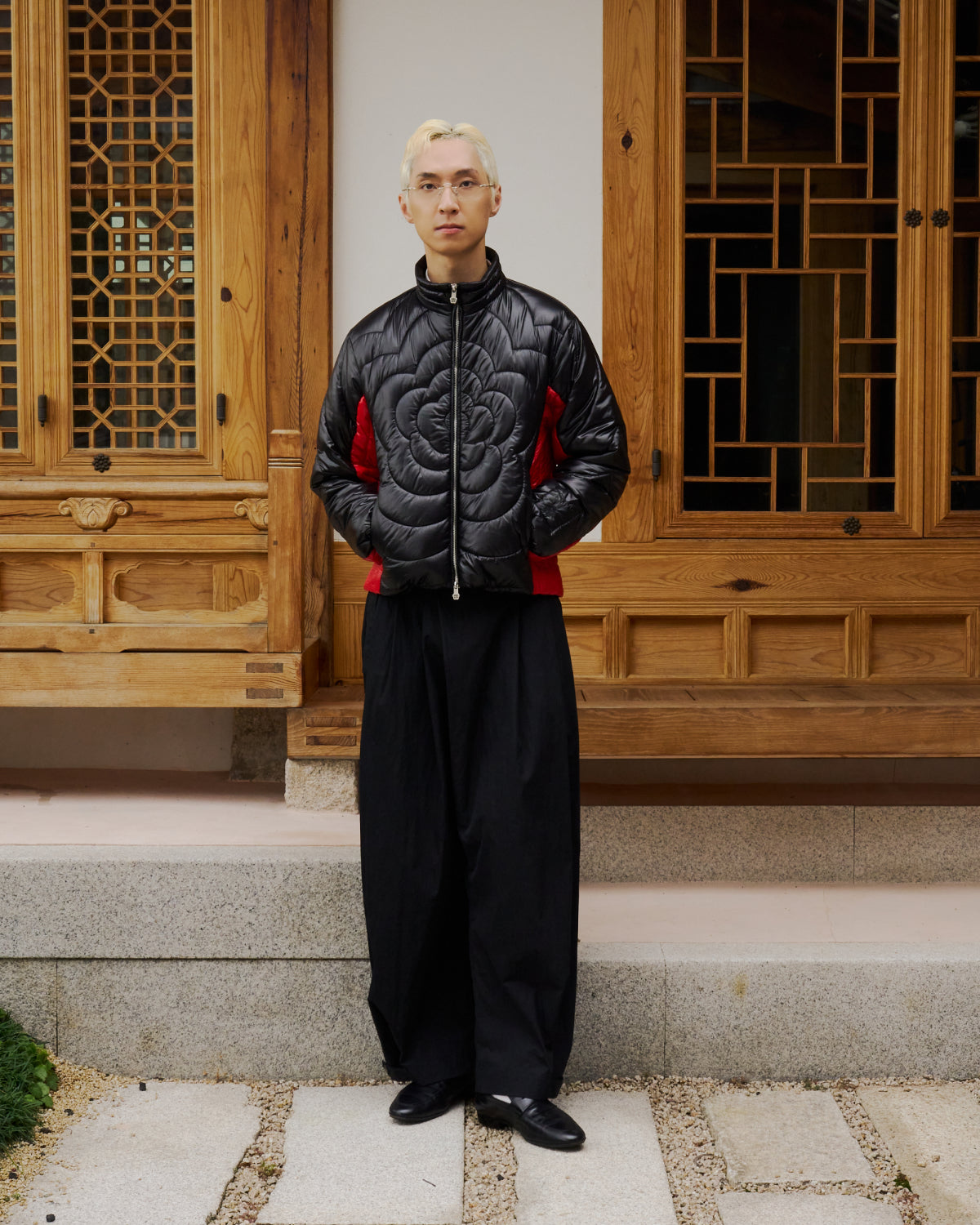 A person with bleach-blond hair and glasses wears the Sundae School BLACK RED MOCHI SPIRAL PUFFER, wide black pants, and black shoes, standing in front of a wooden building with decorative windows.