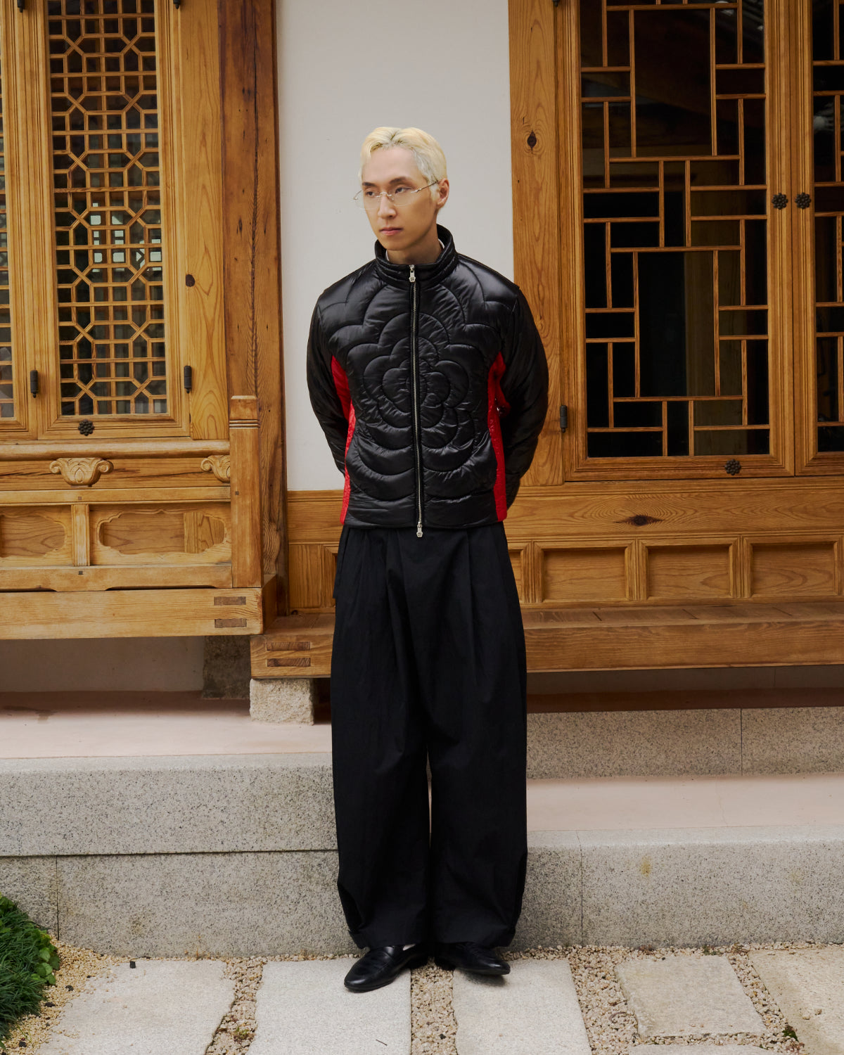 A person with blond hair and glasses wears Sundae School's BLACK RED MOCHI SPIRAL PUFFER, wide black trousers, and black shoes, standing hands behind their back before a traditional wooden building.