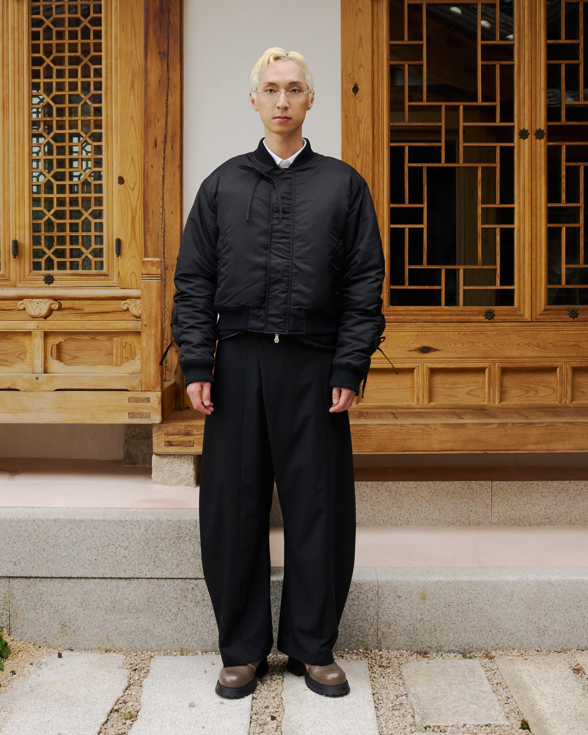 A person with short blond hair, wearing Sundae School's BLACK FLOWER CHANNELED BOMBER, wide black pants, and brown shoes stands on stone steps before a traditional wooden building with detailed window designs.