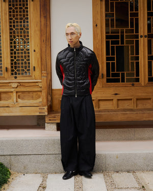 A person with blond hair and glasses wears Sundae School's BLACK RED MOCHI SPIRAL PUFFER, wide black trousers, and black shoes, standing hands behind their back before a traditional wooden building.