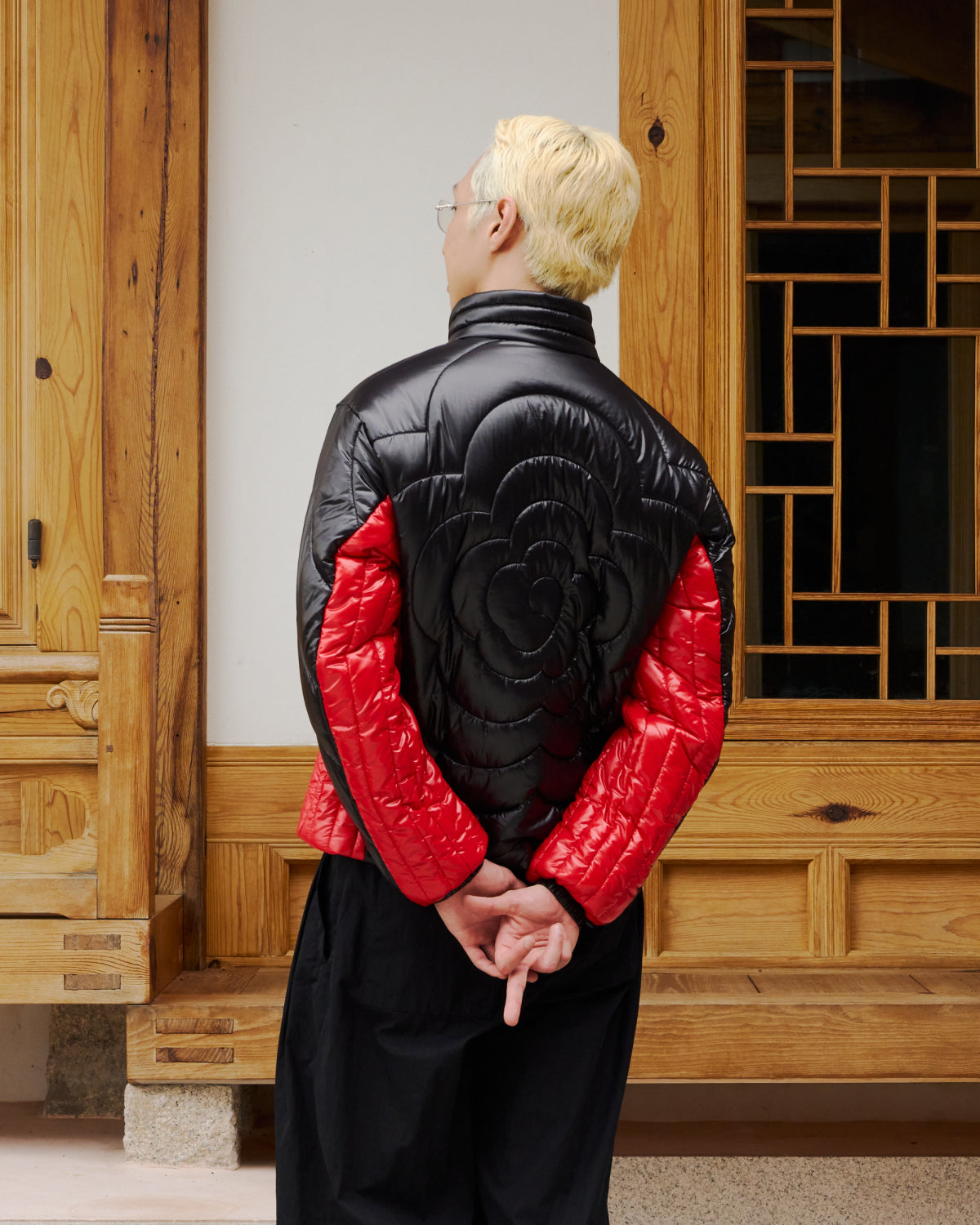 A person with short blond hair and glasses stands with their back to the camera, hands behind them, wearing the Sundae School BLACK RED MOCHI SPIRAL PUFFER in front of a carved wooden door.