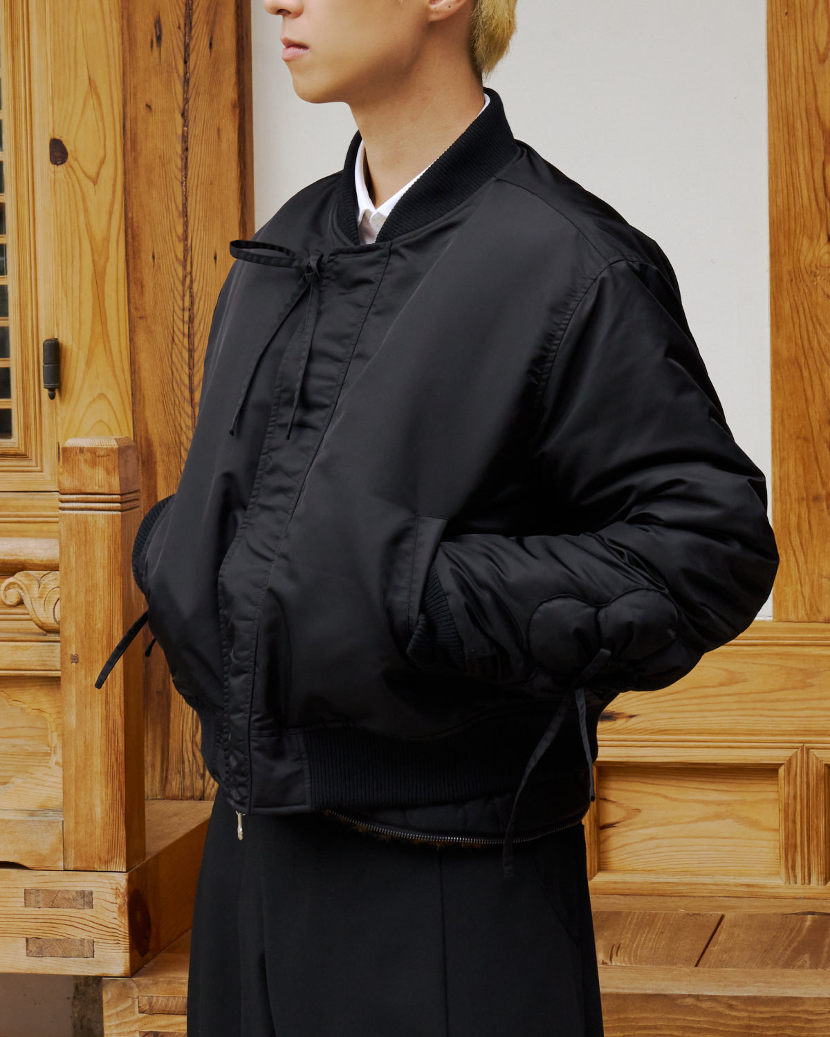 A person with short blond hair wears the BLACK FLOWER CHANNELED BOMBER by Sundae School, standing before a paneled wooden wall. Their face is turned to the side and partially out of frame.