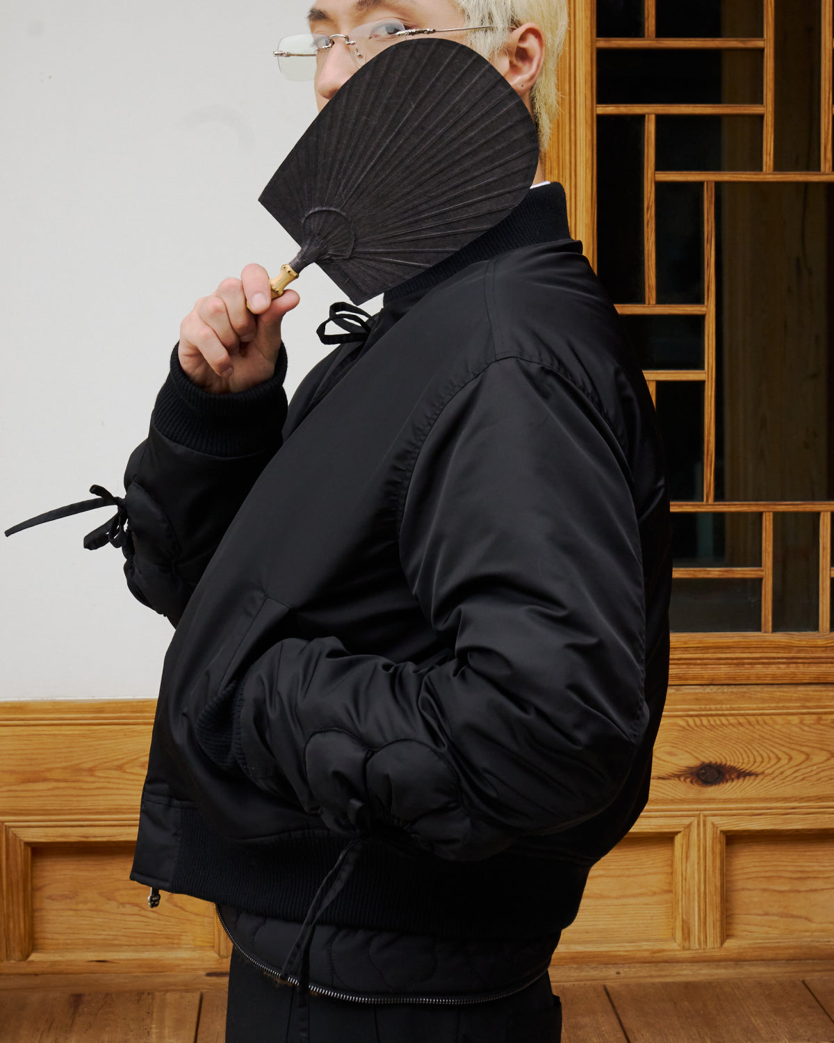 A person with light hair and glasses wears the BLACK FLOWER CHANNELED BOMBER by Sundae School, holding a black fan that partially covers their face, standing indoors before a wooden wall and a geometric grid window.