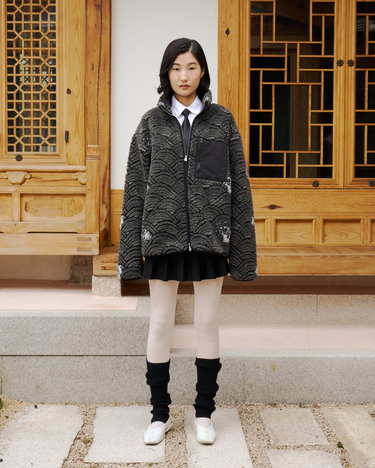 A young woman in front of a traditional wooden building wears Sundae School's BLACK WAVE FLEECE—a textured gray zip-up jacket—styled with a white collared shirt, black skirt, white tights, black leg warmers, and white shoes.