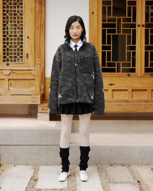 A young woman in front of a traditional wooden building wears Sundae School's BLACK WAVE FLEECE—a textured gray zip-up jacket—styled with a white collared shirt, black skirt, white tights, black leg warmers, and white shoes.