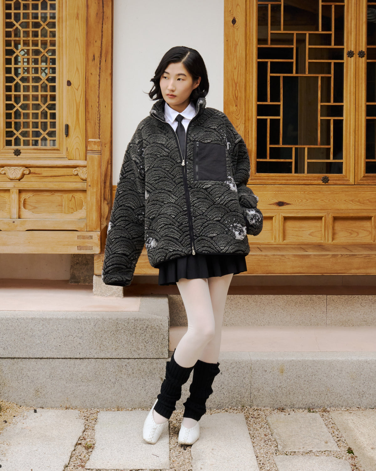 A person with medium-length dark hair wears Sundae School's BLACK WAVE FLEECE, plus a white shirt, black tie, short black skirt, white tights, black leg warmers, and white shoes in front of a traditional wooden building.