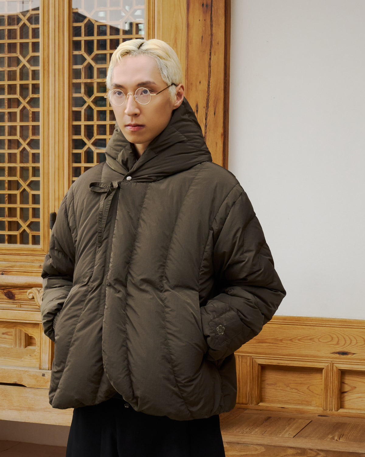 A person with short, bleached blond hair and round glasses wears the Sundae School KHAKI HEAVYWEIGHT MOCHI PUFFER, standing in front of a wooden building with intricate window designs.