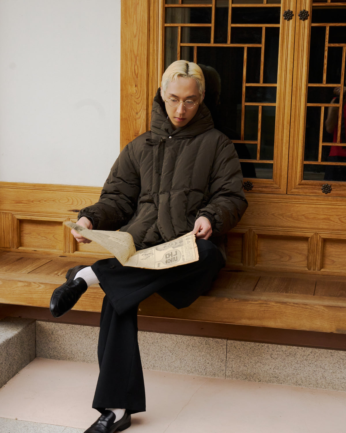A person with blond hair and glasses, dressed in Sundae School’s Khaki Heavyweight Mochi Puffer and black pants, sits on a wooden bench reading a newspaper before a geometric-patterned wooden door.