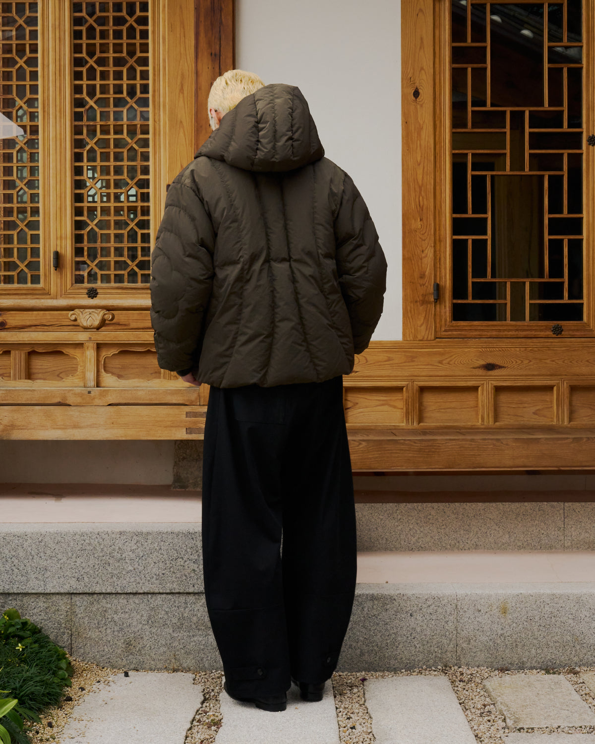 Wearing the KHAKI HEAVYWEIGHT MOCHI PUFFER from Sundae School and wide black pants, a blond-haired individual stands with their back to the camera before a traditional wooden building, channeling a subtle Korean hanbok-inspired look.