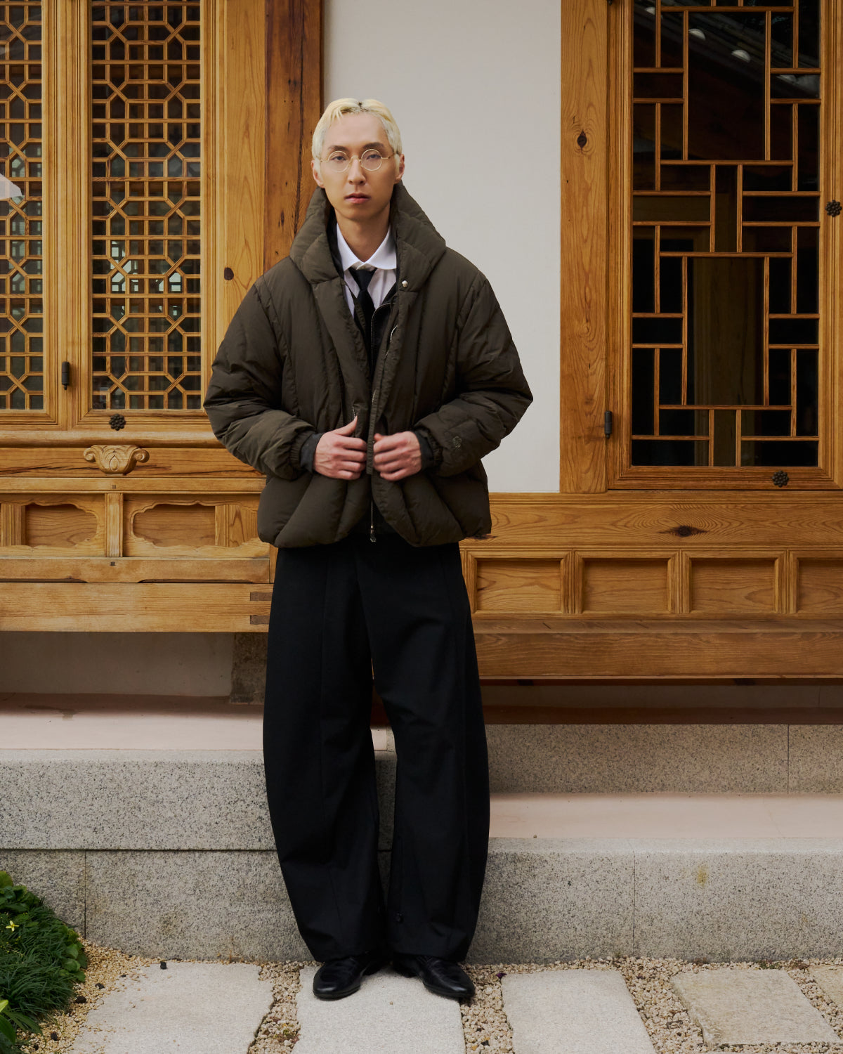 A person with blonde hair, glasses, and a serious expression stands on stone steps in front of a wooden building, wearing Sundae School’s KHAKI HEAVYWEIGHT MOCHI PUFFER jacket, a white shirt, black tie, wide black pants, and black shoes.