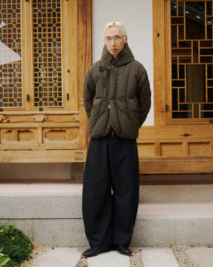 A person with short blond hair and glasses wears the Sundae School KHAKI HEAVYWEIGHT MOCHI PUFFER, wide black pants, and black shoes while standing on stone steps before a traditional wooden building.
