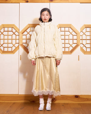 A woman stands indoors against a wooden wall with geometric cutouts, wearing the Sundae School IVORY PLEATED DUVET PUFFER over a satin dress with lace trim, white socks, heeled shoes, and a delicate netted headpiece.