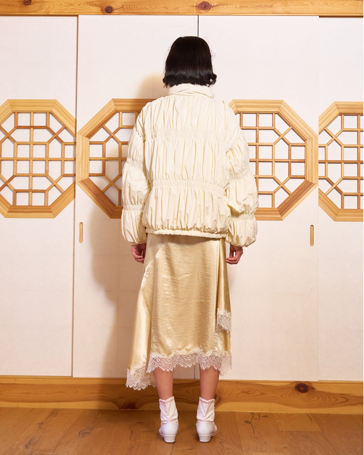A person with short dark hair stands with their back to the camera, wearing the Sundae School IVORY PLEATED DUVET PUFFER, a gold satin lace-trim skirt, and white shoes in front of geometric wooden doors.