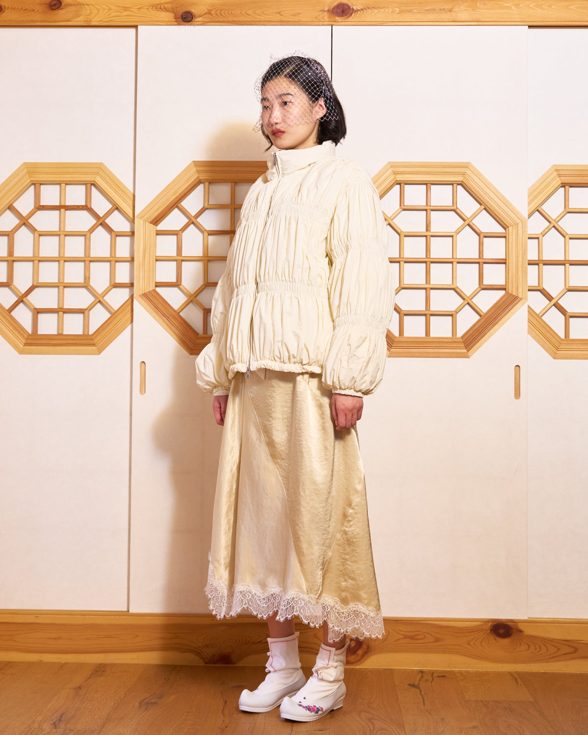 Indoors on a wooden floor before paneled doors, a woman wears the Sundae School IVORY PLEATED DUVET PUFFER, a satin lace-trim skirt, decorative white shoes, and a small netted veil on her head.
