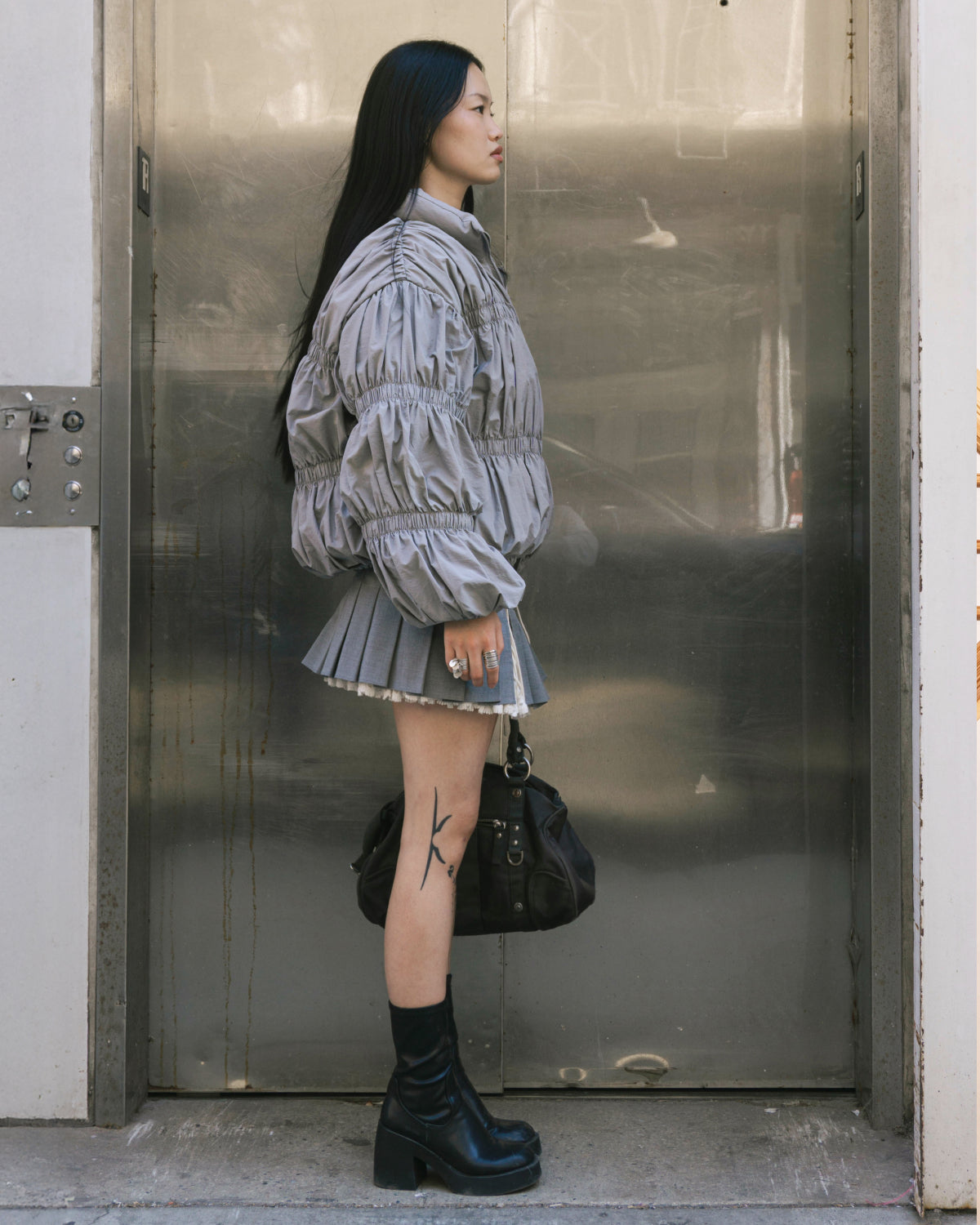 A woman with long black hair and a visible thigh tattoo stands in profile by an elevator, wearing the Sundae School SMOKE PLEATED DUVET PUFFER, black platform boots, and holding a large black bag.