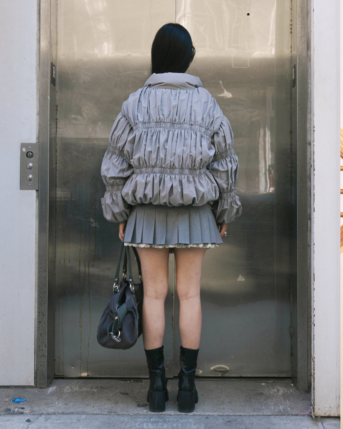 A person with long dark hair and black boots, holding a gray handbag, stands facing an elevator wearing the Sundae School SMOKE PLEATED DUVET PUFFER, a matching gray pleated skirt, and metallic doors reflect their image.