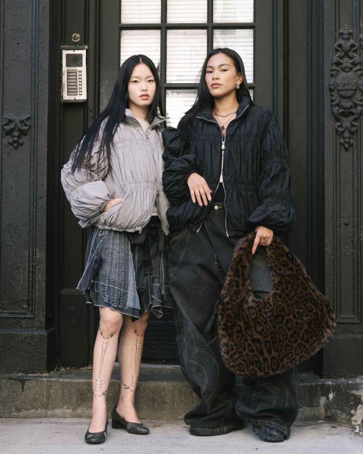 Two women with long dark hair stand before a black door. One wears the Sundae School SMOKE PLEATED DUVET PUFFER, plaid skirt, and flats; the other sports a black jacket, wide-leg jeans, and carries a leopard-print bag. Both look serious.