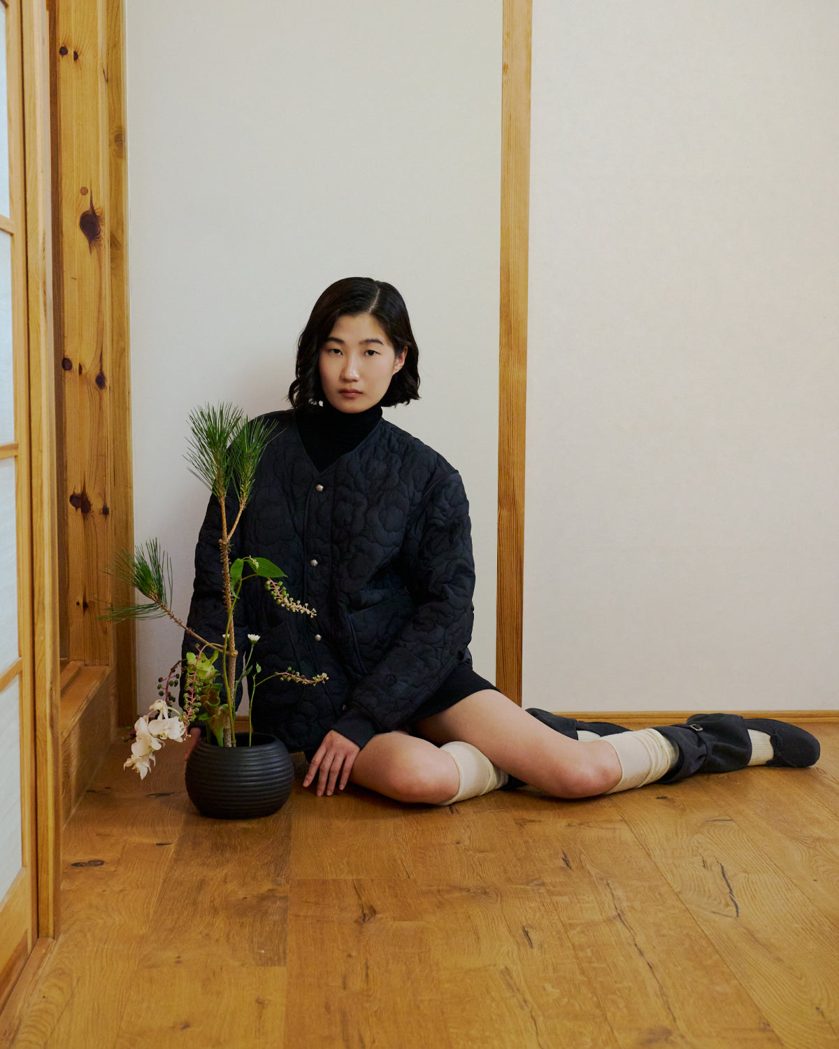 A person with short dark hair sits on a wooden floor in a minimalist room, wearing the Sundae School BLACK MOCHI QUILTED MILITARY LINER jacket, black top, shorts, and leg warmers. A vase with an ikebana floral arrangement is beside them.